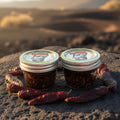 Two jars of Chili Flores hot sauce with chili peppers on a sandy background
